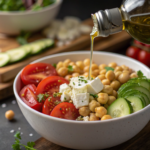 Close-up of a Mediterranean chickpea bowl being drizzled with olive oil dressing, showcasing vibrant ingredients like cherry tomatoes, cucumber, olives, and feta cheese with a blurred background for emphasis.
